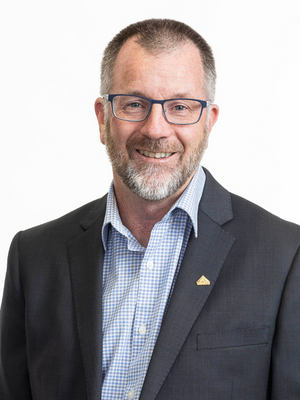 Portrait of a man with short grey hair, glasses and a beard wearing a dark suit jacket and pale blue check shirt