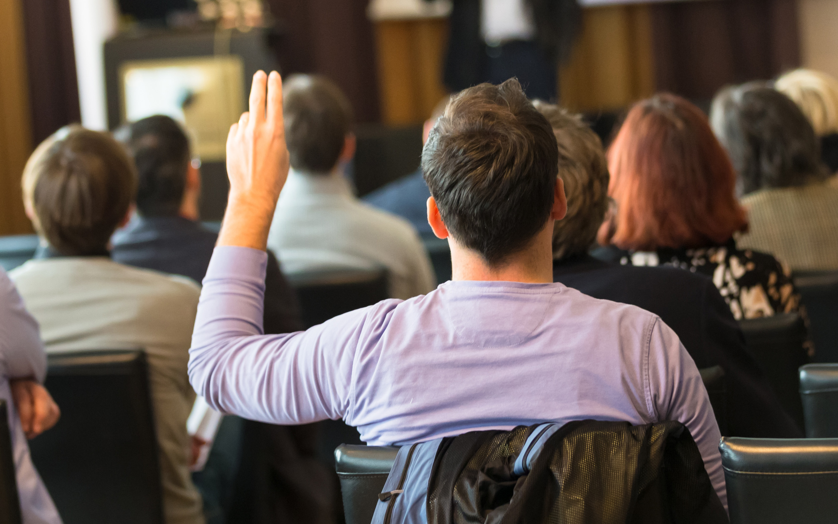 Rear view of a person seated in amongst others with their hand raised